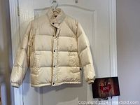 Front view of cream-colored puffy quilted bomber jacket hanging on a white door with snap buttons closed.