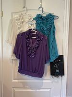Photo showing three feminine ruffle tops hung on hangers on a white door: ivory sheer top with ruffles and buttons, teal sleeveless top with floral ruffles, and purple top with shiny ruffles and elastic waist.