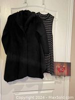 Black sweater coat with hood and button closure hanging on a door, alongside a lightweight sleeveless striped black and white shirt/dress.