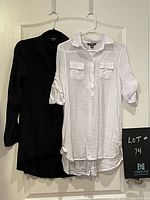 Photo of two long women's shirts hanging on a door: one white with 3/4 sleeves and front pockets, one black with long sleeves and collar.