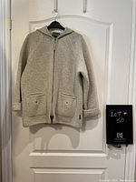 Front view of the grey wool hooded zip-up sweater hanging on a black hanger, showing full length and pockets with embroidered star designs.