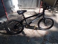 Full side view of the black Mongoose BMX bike leaning against a hockey net outdoors, showing the bike's entire frame, wheels, seat and handlebars.
