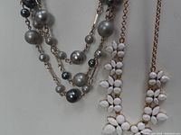 Close-up of grey faux pearl multi-strand necklace with gold-tone leaf accents and part of white flower cluster necklace.