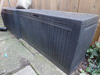 Dark grey Keter resin deck box shown from an angled side view outdoors on a walkway beside a wooden fence. Surface shows dirt and debris from outdoor exposure.