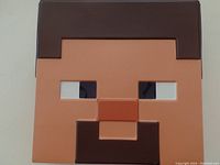 Close-up photo of Minecraft mask showing blocky pixel face with brown hair, eyes, nose, and mouth