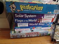 Box view of multiple games and puzzles stacked, showing titles: Perfection, Solar System 500-piece puzzle, Flags of the World 500-piece puzzle, World Map 500-piece puzzle, The Human Body puzzle, Monopoly City game.