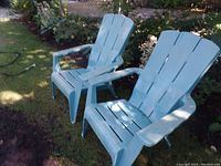 Two mint green plastic Adirondack chairs placed on a lawn near garden plants and a fence.