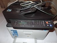 Front and top view of black Samsung multifunction laser printer with control panel and cords on top.