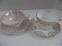 Two bone china teacup and saucer sets with gold patterns, one with a dense leaf pattern and scalloped edges, the other with a floral garland pattern and smooth edges