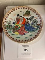 Front view of the antique Famille Rose porcelain plate showing four traditional Chinese figures seated in a natural landscape with birds, rocks, and trees. The border has an intricate red scrolling pattern.