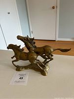 Front angled view of bronze sculpture showing two galloping horses on a base, placed on a white surface inside a room.