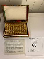 Gold-colored Chinese abacus paperweight inside a decorative box with multicolor floral interior and padded lid.