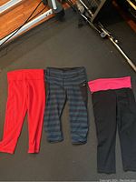Three pairs of Lululemon yoga pants laid out on floor: solid red, black with blue horizontal stripes and pocket, black with wide pink waistband.