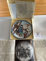 Porcelain plate in box with traditional multi-color painting of a Chinese woman at an indoor desk, accompanied by a literature sheet.