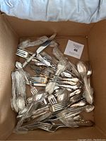 Photo of a cardboard box filled with various silver cutlery including forks, spoons, and knives, some wrapped in plastic.