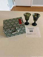 Two natural black and green jade miniature wine glasses or goblets positioned next to their original fabric box with floral geometric pattern on top.