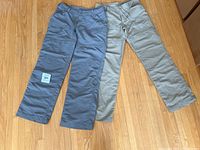 Two pairs of hiking pants laid out flat on wooden floor, showing front view including waist closure and pockets. Left pair is grayish blue, right pair light gray.