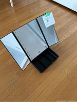 Open view of black tri-fold vanity mirror with lights around the central panel, showing touch power button and divided storage base under the mirrors.