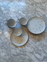 Top-down view of 6 cups stacked inside each other alongside 6 large dessert plates and 6 matching saucers, all white porcelain with gold fleur-de-lis pattern and gold rims.