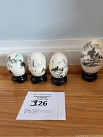 Four vintage hand painted Oriental eggs on black stands showing detailed Chinese landscape scenes placed on wooden floor against baseboard