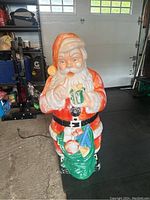 Front view of vintage light up Santa Claus lawn decoration showing Santa holding a gift and green toy sack, paint wear visible