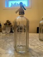 Single vintage Magda soda water spritzer bottle made of clear ribbed glass with metal top and branding on front.