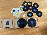 Top-down image of the vinyl lot showing several 45 RPM singles with various record label colors and some paper sleeves alongside three 12-inch vinyl LP records in cardboard sleeves on a wooden floor.