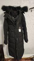 Front view of women's black long winter jacket parka with fur-trimmed hood hanging on hanger against textured wall.