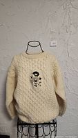 Cream cable knit sweater with sheep embroidery on front, displayed on wire mannequin against textured white wall.