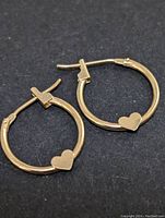 Pair of 14K gold hoop earrings with small heart detail, shown lying on black background.