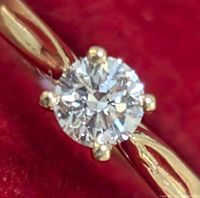 Close-up image of the round brilliant cut lab-created diamond set in 10K yellow gold four-prong setting on the ring band against a red background.