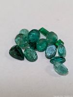 Twelve loose polished emerald gemstones on white background showing various cuts and shades of green.