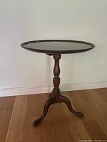 Side view of antique wooden side table showing the turned pedestal and tripod curved legs on a wooden floor.
