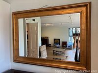 Front-facing photo of a large rectangular wall mirror with a wide gold frame hung on a white wall. The mirror reflects an interior room with furniture and windows.