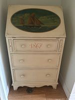 Front view of cream-colored fold-out desk with oval nautical painting of ships and '1867' on top drawer.