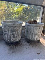 Two vintage outdoor planters with raised floral patterns on their sides, placed on a concrete floor near a window with trees outside.