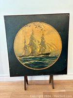 Front view of square folding table upright showing painted sailing ship image on table surface with some wear.