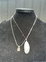 Two sterling silver necklaces displayed on a black mannequin bust, one with small square amber pendant and one with large agate slice pendant.