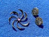 Photo of pinwheel brooch with central amethyst cabochon alongside pair of swirled abalone sterling silver screwback earrings on blue background.