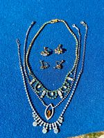 Full view of three vintage rhinestone necklaces and two pairs of earrings displayed on blue background