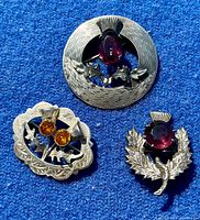 Three vintage Scottish sterling silver brooches with thistle motifs and colored gemstones on blue fabric background