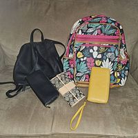 Photo shows two backpacks, one black leather and one floral pattern, along with three wallets arranged on a couch.