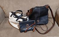 Three leather bags grouped on a beige couch: white handbag, black crossbody purse with brown accents, and black clutch wallet with tan tassel wrist strap.