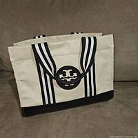 Front view of tote showing cream canvas body, black leather base, striped black and white handles, and circular black leather logo.