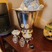 Silver toned champagne bucket with attached stand and decorative feet, shown with glass and silver cruet set and silver serving platter.