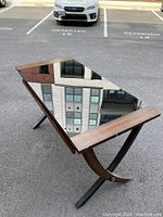 Rectangular glass top desk with wood side framing and black metal curved base support, photographed outdoors on asphalt near parked car.