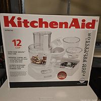 Front of box showing KitchenAid branding, 12-cup capacity, ultra wide mouth feed tube, and images of included bowls and accessories.