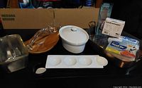 Photo showing various kitchen items including glass cake server, white ceramic casserole with lid, metal loaf pan, white ceramic egg dish, and copper casserole in background