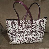 Front and side view of the leather tote bag with cream and maroon leaf pattern and maroon leather handles placed on a couch.