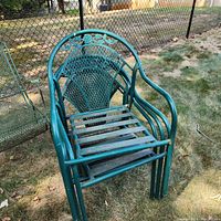 Four green metal outdoor chairs stacked on grass near a chain link fence. Chairs have curved armrests and decorative mesh backs.